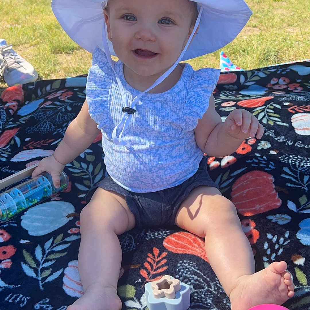 Ella is registered to the contest to win money with this photo: baby, clothing, face, fun, grass, hat, head, leisureactivities, nature, outdoors, park, person, photography, picnic, plant, portrait, shorts, sitting, summer, sunhat