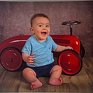 Thélio participe au concours pour gagner de l'argent avec cette photo : baby, child, toddler, toy_car, red, blue_shirt, shorts, barefoot, floor, wooden_floor, studio, portrait, smiling, happy, indoor, sitting, playful, steering_wheel, background, vintage_toy