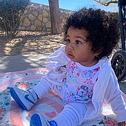 Amira is registered to the contest to win money with this photo: baby, blanket, blue_shoes, child, curly_hair, cute, expression, footwear, garden, infant, nike, outdoor, playtime, side_view, sitting, stone_wall, sunny, toddler, tree, white_clothing