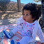 baby, child, outdoor, curly_hair, white_clothing, blue_shoes, nike, blanket, sitting, garden, tree, stone_wall, sunny, side_view, cute, infant, toddler, footwear, expression, playtime
