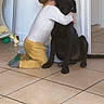 child, dog, hug, black_dog, floor, tile_floor, indoors, doorway, affection, friendship, yellow_pants, long_sleeve_shirt, kneeling, pet, love, companion, home, cute, animal, tiled_floor