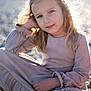 child, girl, blonde_hair, long_hair, dress, beach, pebbles, sunlight, backlit, portrait, smile, sitting, casual_pose, outdoor, nature, soft_light, young, happy, relaxed, daytime