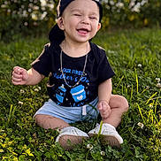 Avery is registered to the contest to win money with this photo: baby, child, grass, outdoor, smiling, happy, cap, black_tshirt, shorts, white_shoes, necklace, nature, sunlight, greenery, cute, sitting, portrait, daylight, casual_clothing, joyful