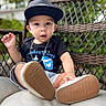 toddler, child, cap, black_shirt, mother's_day, chain_necklace, shorts, sneakers, shoe_sole, wicker_chair, cushion, outdoor, greenery, portrait, curious_expression, casual_clothing, baby, seated, daylight, closeup