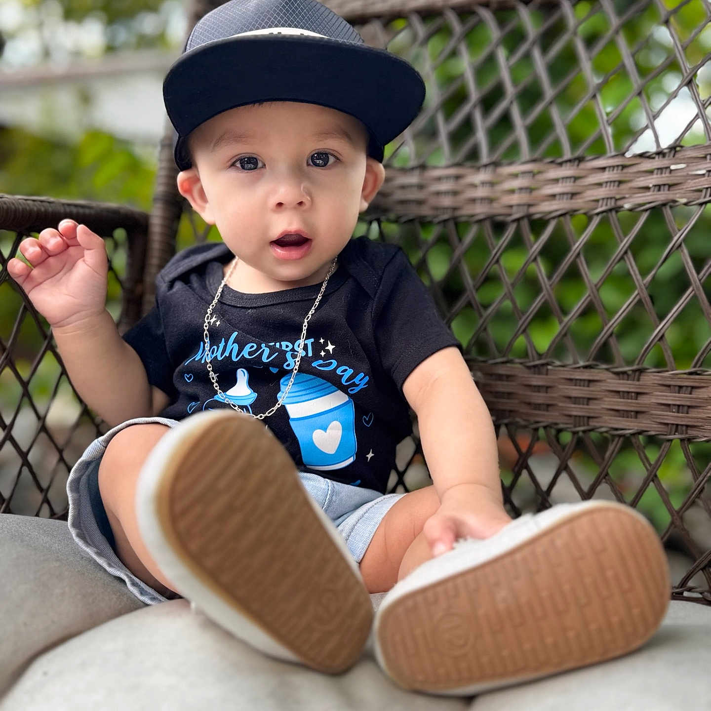 Avery is registered to the contest to win money with this photo: toddler, child, cap, black_shirt, mother's_day, chain_necklace, shorts, sneakers, shoe_sole, wicker_chair, cushion, outdoor, greenery, portrait, curious_expression, casual_clothing, baby, seated, daylight, closeup