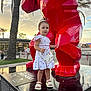 Naïlya participe au concours pour gagner de l'argent avec cette photo : child, curious, daylight, dress, footwear, girl, outdoor, palm_trees, people, portrait, public_space, purse, red_sculpture, sandals, standing, statue, sunset, toddler, urban, white_dress