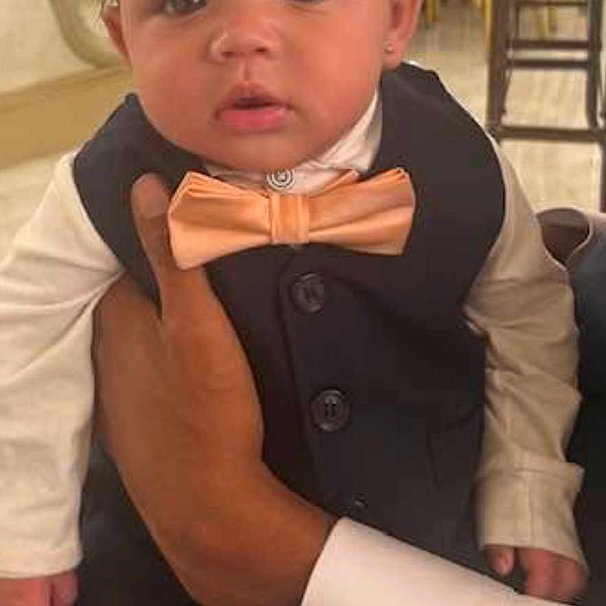 Ali is registered to the contest to win money with this photo: baby, child, tuxedo, bow_tie, hand, indoor, formal_wear, person, face, portrait, curly_hair, earrings, table, chair, dining_room, adult_hand, curious_expression, suit, white_shirt, event