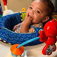Ali joined the competition — help win amazing prizes! baby, infant, child, fingers_in_mouth, baby_activity_center, toy, red_toy, blue_seat, indoor, curly_hair, cute, playtime, closeup, expression, young_child, seat, colorful, rattle, adorable, portrait