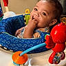 baby, infant, child, fingers_in_mouth, baby_activity_center, toy, red_toy, blue_seat, indoor, curly_hair, cute, playtime, closeup, expression, young_child, seat, colorful, rattle, adorable, portrait