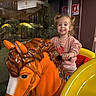 child, toddler, smiling, pigtails, carousel_horse, orange_horse, toy, ride, yellow, pink_clothing, indoor, window, nighttime, happy, person, play, amusement, fun, grip, hair_ties