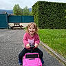 child, toddler, toy_car, pink, outdoor, smiling, happy, sweater, grass, hedge, pavement, picnic_table, vehicle, play, person, casual_clothing, nature, daylight, fun, small_vehicle