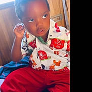 Shakur White is registered to the contest to win money with this photo: child, toddler, shirt, red_pants, indoors, wooden_wall, curious_expression, seated, colorful_clothing, young_child, person, portrait, casual_wear, farm_print, short_hair, hand_near_face, baby, cute, cozy, natural_light