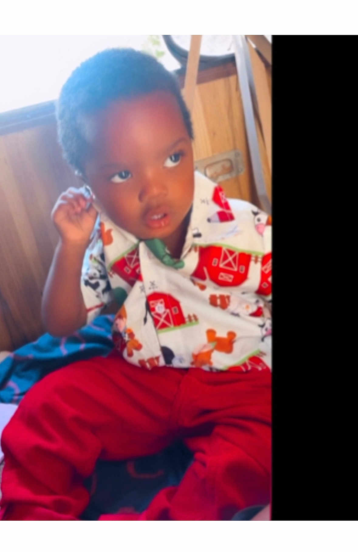 Shakur White is registered to the contest to win money with this photo: child, toddler, shirt, red_pants, indoors, wooden_wall, curious_expression, seated, colorful_clothing, young_child, person, portrait, casual_wear, farm_print, short_hair, hand_near_face, baby, cute, cozy, natural_light