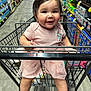 baby, shopping_cart, grocery_store, aisle, smile, child, pink_dress, happy, face, infant, legs, metal_cart, floor, shelf, products, beverages, clothing, cute, indoor, holding