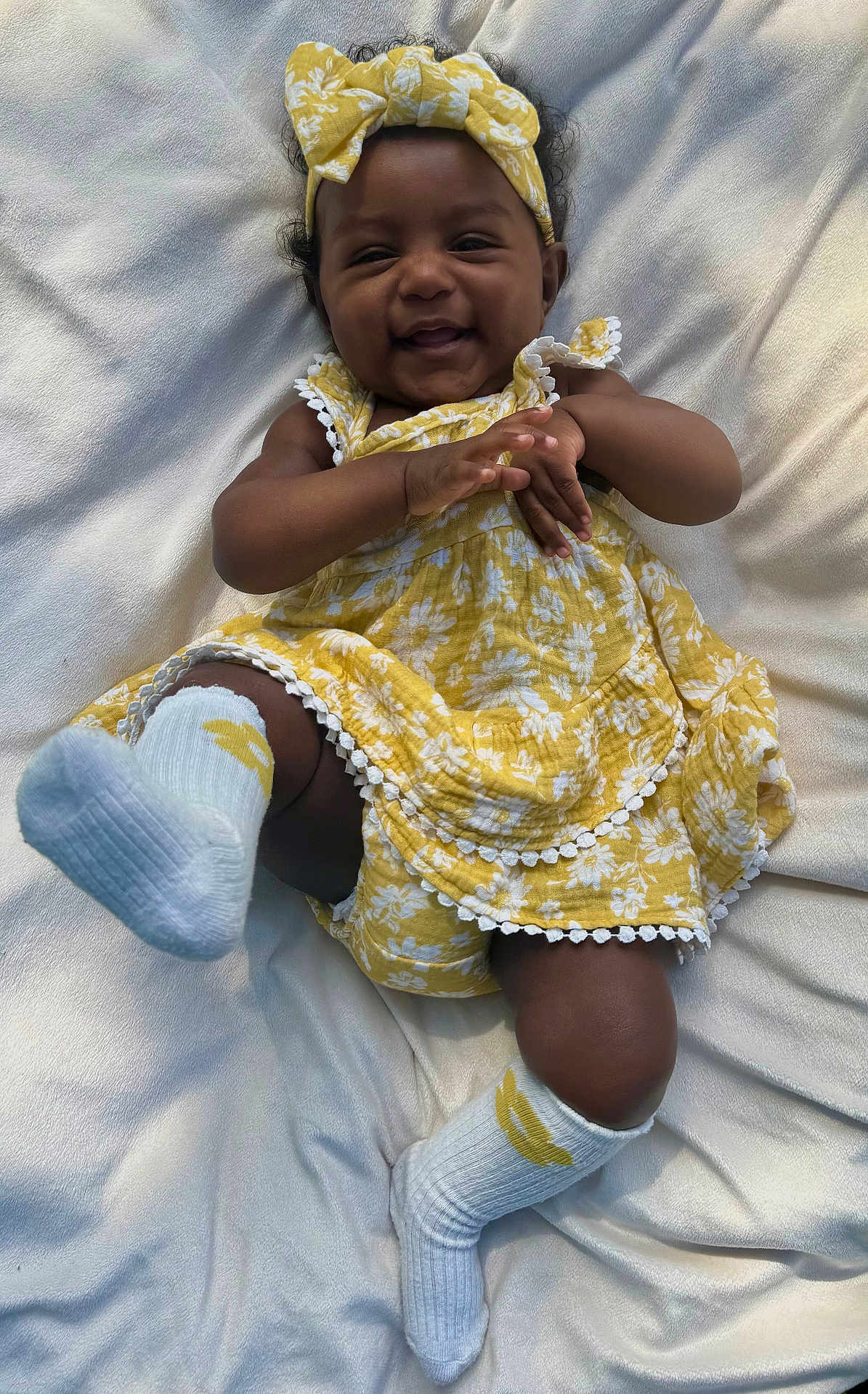 Nayvee is registered to the contest to win money with this photo: baby, child, smiling, yellow_dress, headband, white_blanket, sock, happy, infant, cute, lying_down, playful, floral_pattern, toddler, one_person, indoors, skin, face, hands, feet