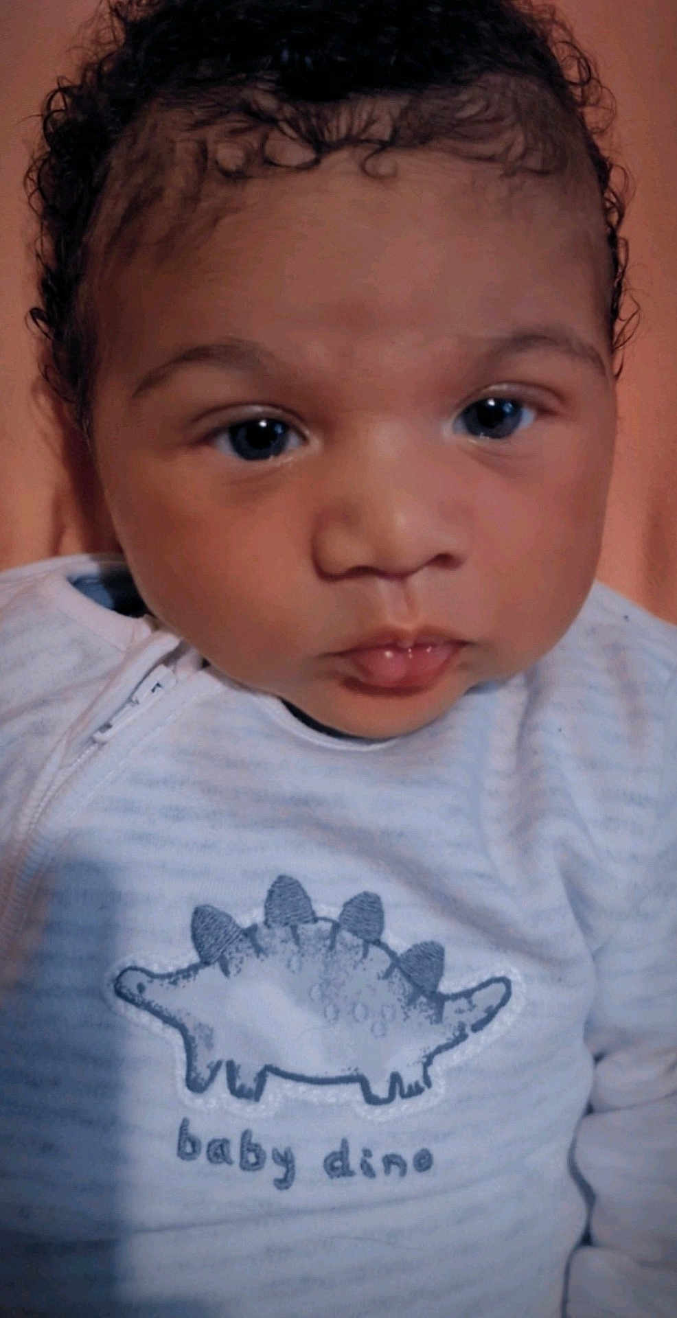 Ruben is registered to the contest to win money with this photo: baby, infant, face, eyes, curly_hair, clothing, onesie, dinosaur_graphic, baby_dino, portrait, close_up, cute, child, young, soft_lighting, indoor, skin, mouth, nose, expression