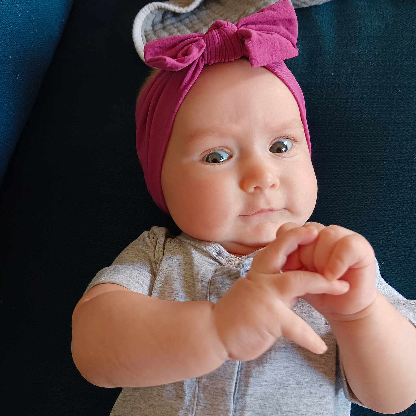 Stella Rose joined the competition — help win amazing prizes! baby, blanket, child, closeup, clothing, couch, curious, cute, expression, face, gray_onesie, hands, headband, indoor, infant, person, pink_bow, portrait, skin, soft_lighting