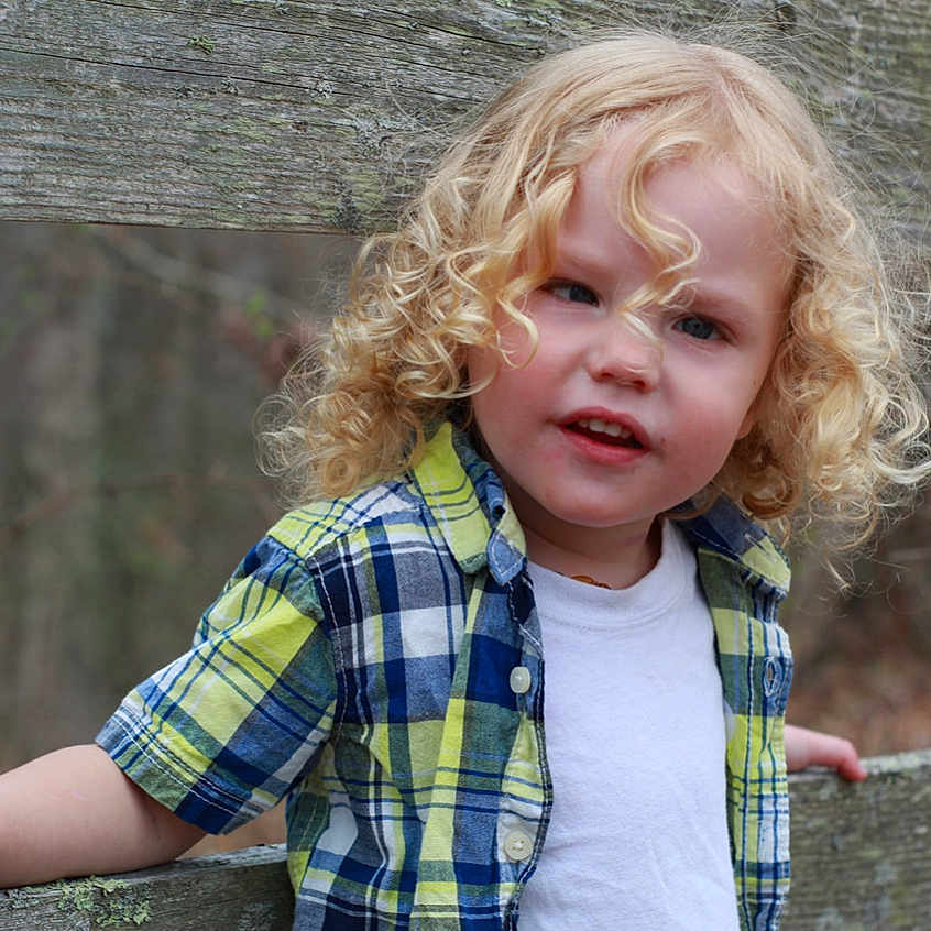 Caliber joined the competition — help win amazing prizes! blond_hair, casual, child, curly, curly_hair, daylight, expression, forest_background, leaning, nature, outdoor, person, plaid_shirt, playful, portrait, smiling, toddler, white_tee, wooden_fence, young_child