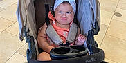 Avia is registered to the contest to win money with this photo: baby, stroller, infant, headband, orange_dress, pouty_face, indoor, tiled_floor, person, child, cute, seat_belt, baby_seat, foot, hand, toy_holder, evenflo, blurred_person, expression, modern_interior