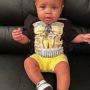 Zacharia is registered to the contest to win money with this photo: toddler, child, yellow_bows, curly_hair, black_tshirt, graphic_tee, yellow_shorts, sneakers, black_leather_couch, sitting, indoors, portrait, cute, young_child, fashion, casual_clothing, person, happy, smiling, baby