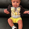 toddler, child, yellow_bows, curly_hair, black_tshirt, graphic_tee, yellow_shorts, sneakers, black_leather_couch, sitting, indoors, portrait, cute, young_child, fashion, casual_clothing, person, happy, smiling, baby