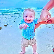 Weston is registered to the contest to win money with this photo: toddler, child, beach, ocean, sand, smiling, holding_hand, blue_shirt, shorts, barefoot, water, sky, waves, person, happy, outdoor, summer, playing, family, vacation