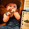Weston is registered to the contest to win money with this photo: baby, child, cowboy_hat, denim_overalls, food, meat, table, menu, restaurant, smiling, cute, indoor, person, appetite, chubby_cheeks, wood, clothing, happy, eating, portrait