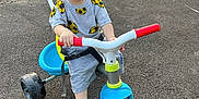 Mehmetcan a rejoint le concours — aidez-le/la à gagner de superbes lots ! toddler, child, tricycle, outdoor, pavement, toy, vehicle, barefoot, curious, blue_eyes, short_hair, gray_clothing, red_handlebars, plastic, playtime, daylight, road, fence, car, urban