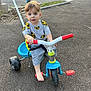 toddler, child, tricycle, outdoor, pavement, toy, vehicle, barefoot, curious, blue_eyes, short_hair, gray_clothing, red_handlebars, plastic, playtime, daylight, road, fence, car, urban