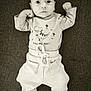 baby, infant, child, dinosaur_graphic, clothing, pants, socks, lying_down, monochrome, black_and_white, cute, portrait, serious_face, raised_fists, text_on_clothing, soft_fabric, indoor, comfort, small_person, casual_wear