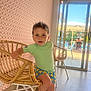 child, boy, indoor, rattan_chair, shorts, patterned_clothing, spiky_hair, window, sliding_door, outdoor_view, pool_area, sunlight, floor, wallpaper, geometric_pattern, furniture, seated, casual, expression, curious