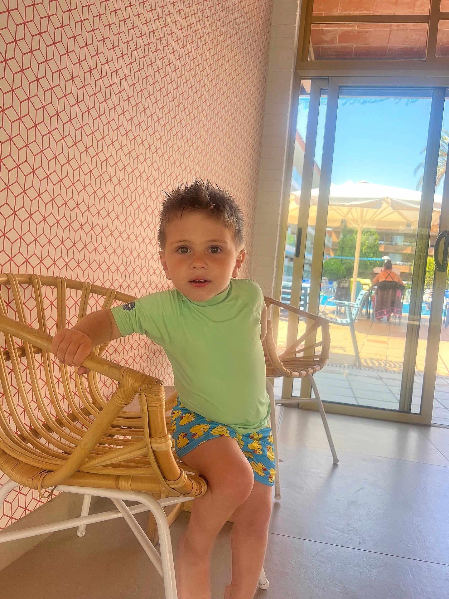 Amelio participe au concours pour gagner de l'argent avec cette photo : child, boy, indoor, rattan_chair, shorts, patterned_clothing, spiky_hair, window, sliding_door, outdoor_view, pool_area, sunlight, floor, wallpaper, geometric_pattern, furniture, seated, casual, expression, curious