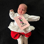Djennah a rejoint le concours — aidez-le/la à gagner de superbes lots ! baby, infant, one_month, clothing, white_top, red_pants, frilly_skirt, card, floral_design, lying_down, black_background, cute, celebration, milestone, newborn, soft_booties, baby_face, happy, small_hands, portrait