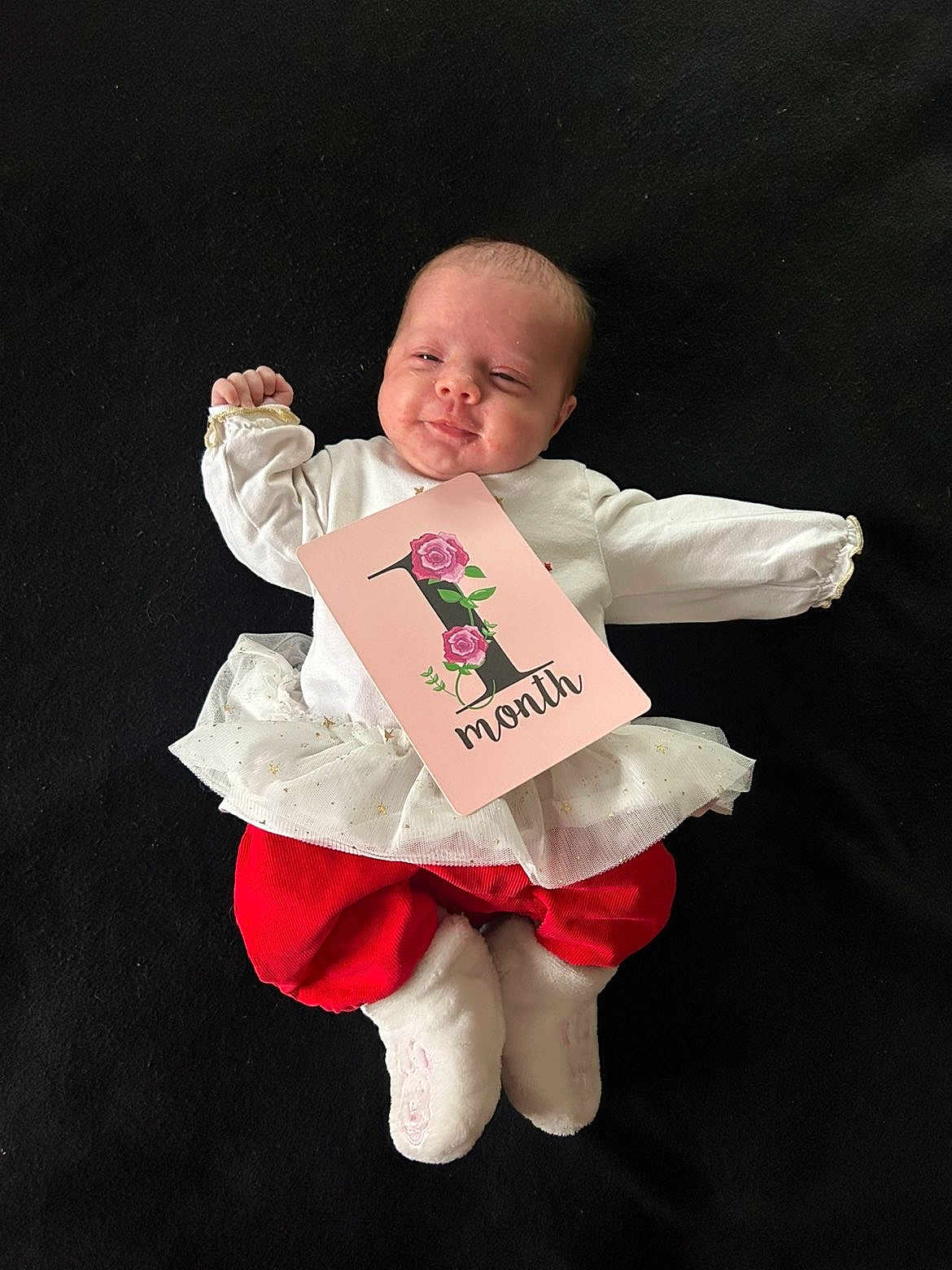 Djennah a rejoint le concours — aidez-le/la à gagner de superbes lots ! baby, infant, one_month, clothing, white_top, red_pants, frilly_skirt, card, floral_design, lying_down, black_background, cute, celebration, milestone, newborn, soft_booties, baby_face, happy, small_hands, portrait