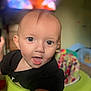 baby, child, face, blue_eyes, curious, indoor, blurred_background, high_chair, black_shirt, infant, tongue_out, closeup, person, home, colorful, toy, seat, expression, young_child, portrait