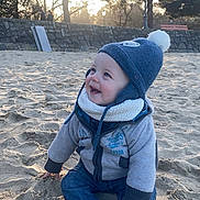 Nolann a rejoint le concours — aidez-le/la à gagner de superbes lots ! baby, child, toddler, beach, sand, winter_hat, coat, jeans, socks, smile, sitting, outdoor, sunlight, trees, stone_wall, lamp_post, bench, portrait, happy, playful