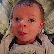 Apollo is registered to the contest to win money with this photo: baby, newborn, infant, face, eyes, tongue_out, onesie, clothing, blanket, bedding, close_up, portrait, expression, cute, skin, hair, cheeks, indoor, lying_down, text_on_clothing