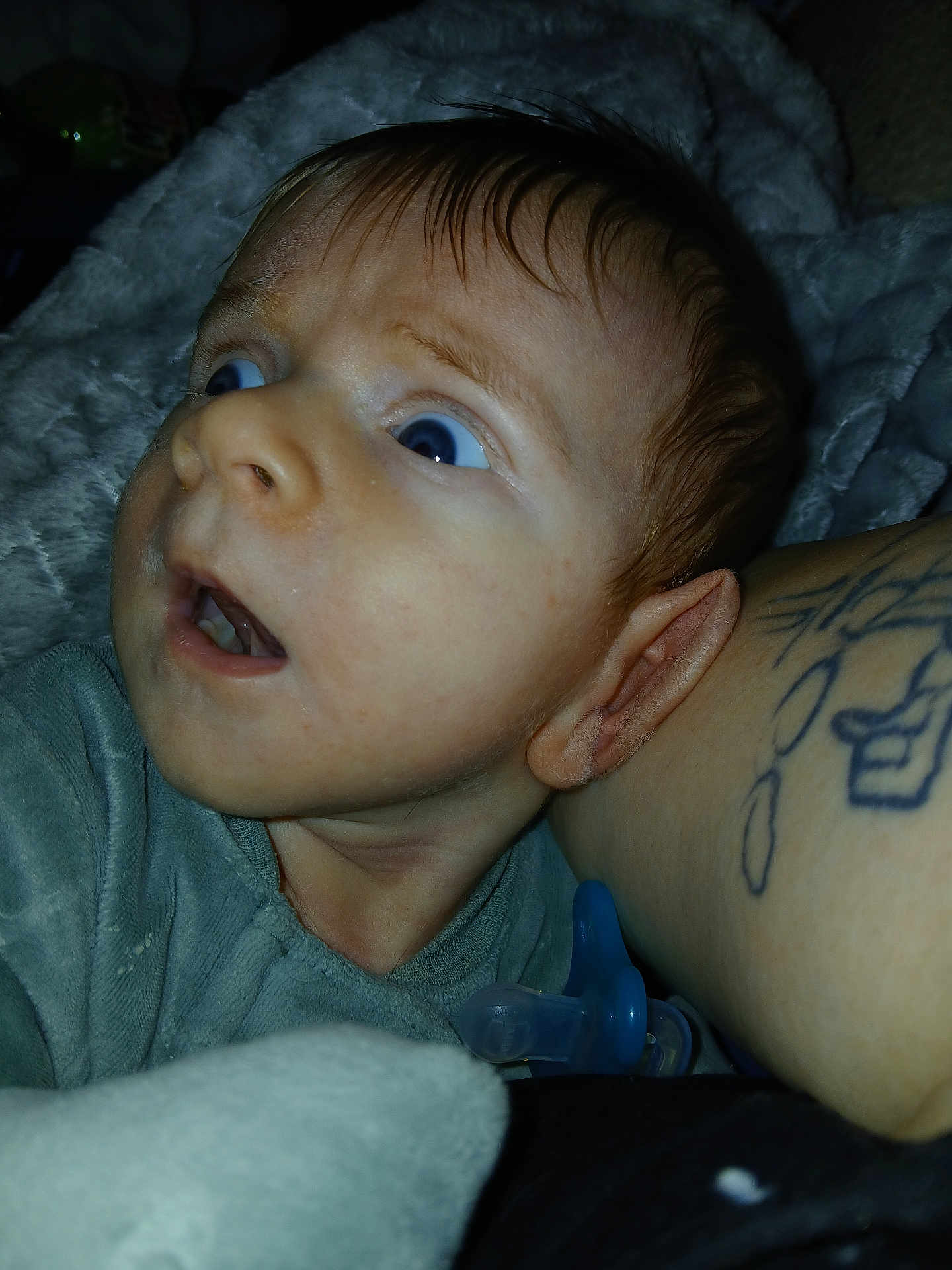 Apollo is registered to the contest to win money with this photo: baby, infant, blue_eyes, surprised_expression, pacifier, tattoo, blanket, close_up, flash_photography, onesie, ear, mouth, skin, parent_arm, indoors, bedtime, drool, portrait, lighting, young_child