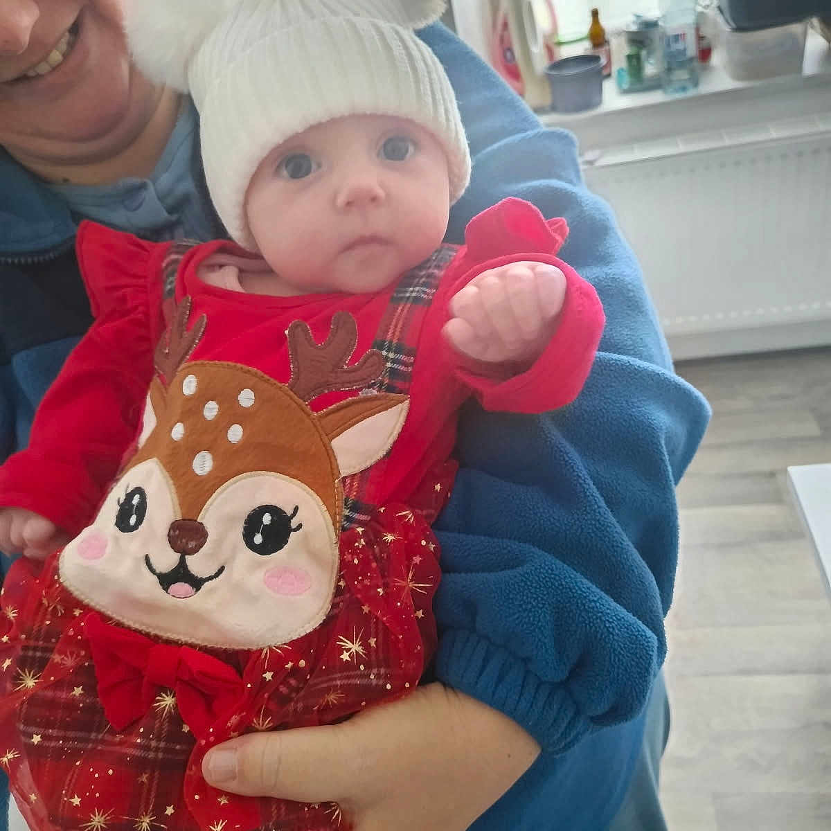 Julia a rejoint le concours — aidez-le/la à gagner de superbes lots ! adult, baby, blue_jacket, child, cute, deer_design, festive, floor, hand, hat, holding, indoor, kitchen, person, plaid, pom_poms, radiator, red_clothing, smile, window