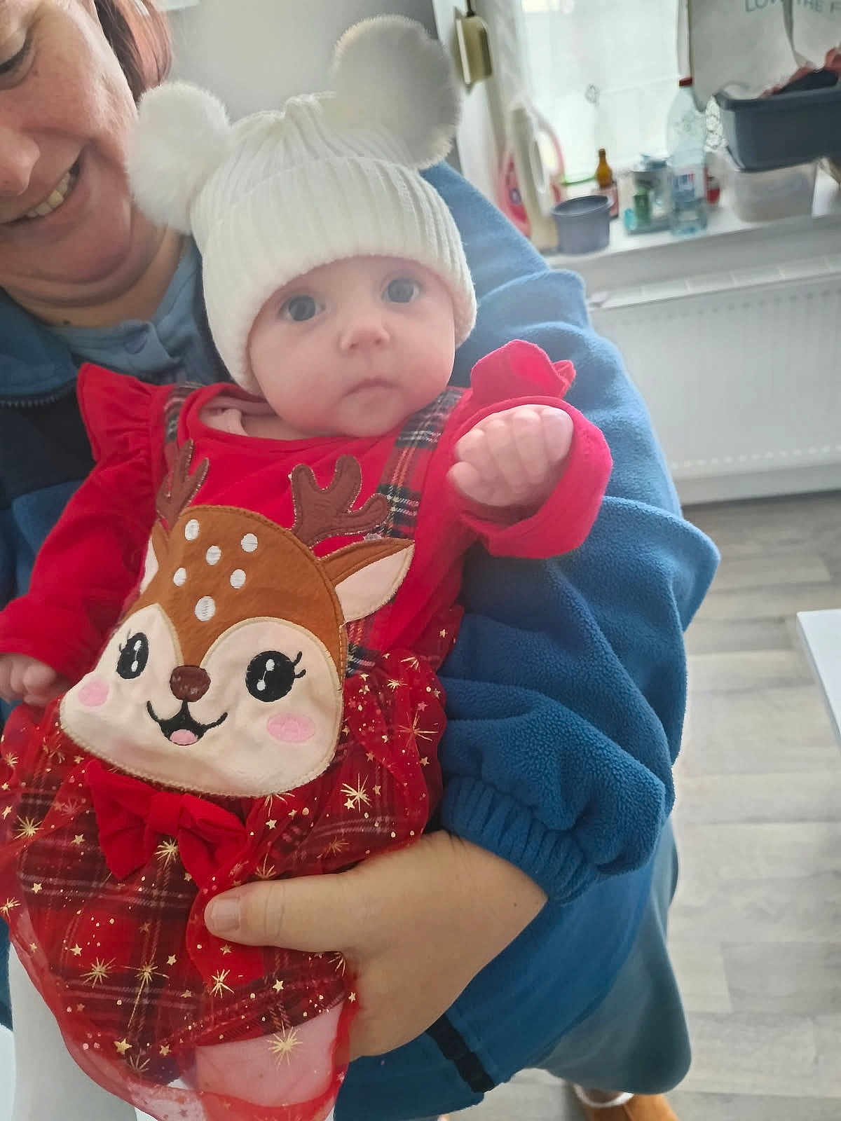 Julia a rejoint le concours — aidez-le/la à gagner de superbes lots ! baby, child, adult, person, hat, pom_poms, red_clothing, plaid, deer_design, smile, hand, indoor, kitchen, window, radiator, floor, blue_jacket, holding, cute, festive