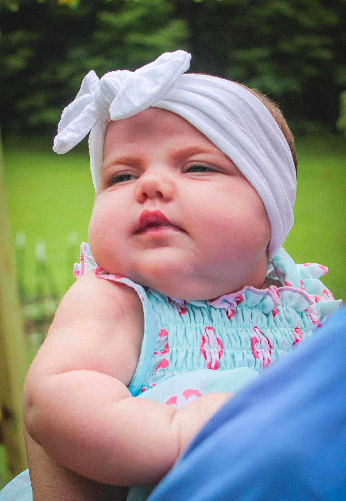 Maddalynn joined the competition — help win amazing prizes! baby, child, headband, bow, outdoor, greenery, pastel_clothing, cute, chubby_cheeks, portrait, soft_skin, infant, holding, person, summer, daylight, closeup, face, expression, young_child