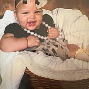 Naiari is registered to the contest to win money with this photo: baby, infant, child, smile, pearl_necklace, headband, bow, black_clothing, wicker_basket, blanket, cozy, portrait, cute, happy, indoor, soft_lighting, fashion_accessories, baby_sock, person, closeup