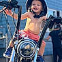 child, toddler, motorcycle, helmet, outdoor, smiling, happy, vehicle, blue_house, person, shoes, shorts, sunlight, metal, handlebars, blurred_background, fun, casual, daytime, playful