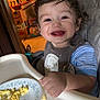 Adrian is registered to the contest to win money with this photo: toddler, child, smiling, high_chair, scrambled_eggs, food, plate, curly_hair, indoor, kitchen, happy, meal, baby, person, furniture, tray, clothing, blue_stripes, face, hand