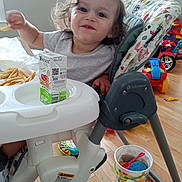 Adrian is registered to the contest to win money with this photo: toddler, child, high_chair, fries, juice_box, smile, curly_hair, indoor, toy, wooden_floor, casual, meal, happy, person, plastic_bucket, red_toy, baby_trend, striped_shirt, window, home