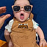 baby, blue_background, button, child, clothing, cute, expression, face, hair, hand, indoor, infant, monster_prints, mouth_open, orange_surface, person, romper, short_sleeves, sitting, sunglasses