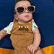 Asher joined the competition — help win amazing prizes! baby, child, sunglasses, romper, brown_clothing, blue_cushion, orange_blanket, indoor, cute, infant, person, portrait, fashion, casual, seated, funny_expression, hair, cute_outfit, young_child, relaxed