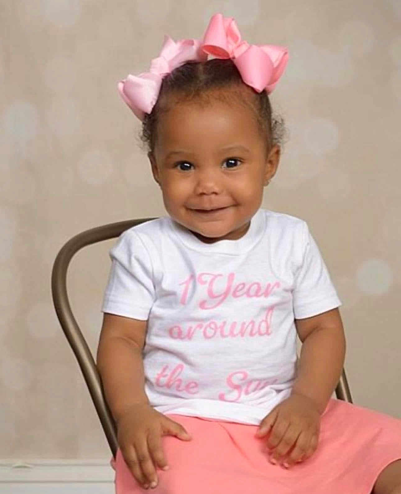Keziah joined the competition — help win amazing prizes! baby, background, chair, child, clothing, cute, face, fashion, girl, hands, happy, indoor, one_year, pink_bow, pink_skirt, portrait, sitting, smile, toddler, white_shirt