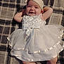 Willow is registered to the contest to win money with this photo: baby, infant, smiling, white_dress, headband, plaid_blanket, lying_down, happy, cute, child, portrait, indoors, fabric, soft, toddler, person, skin, expression, fashion, relaxing