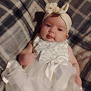 Willow is registered to the contest to win money with this photo: baby, infant, dress, headband, bow, plaid_blanket, lying_down, soft_lighting, cute, child, white_clothing, portrait, indoors, person, toddler, fabric, smiling, face, head, cozy